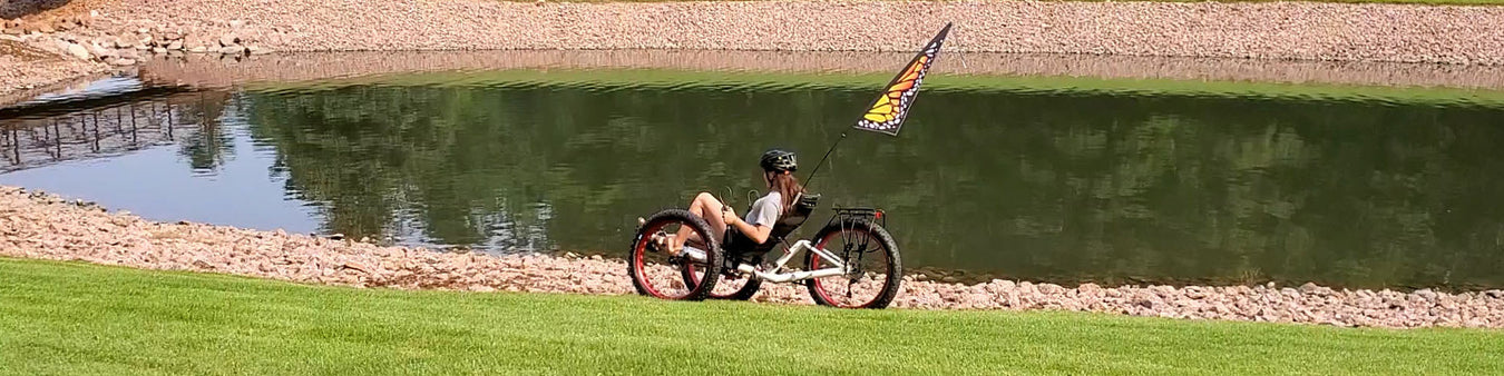 A girl rides a white Azub Fat trike with a Monarch butterfly themed safety flag across the grass next to a rocky shoreline around a pond.