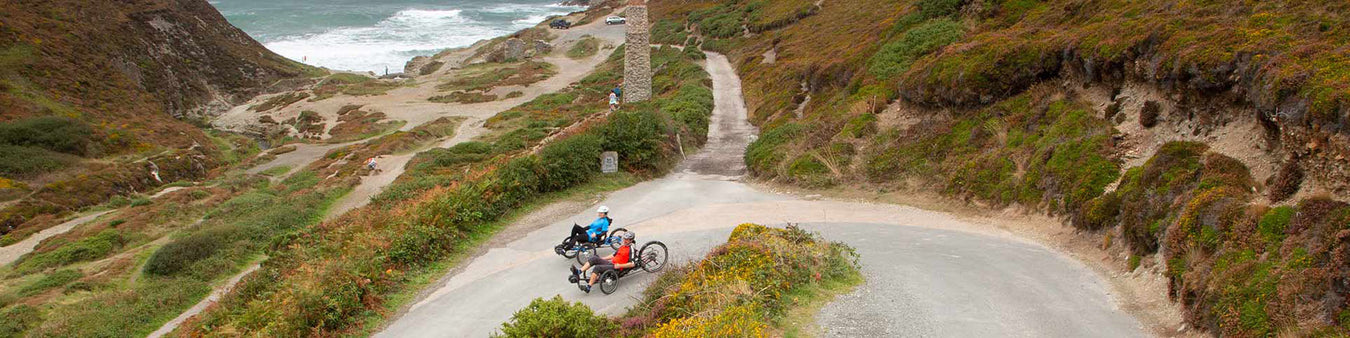 Two people ride recumbent trikes down a switchback of hilly terrain along the coast of Cornwall.