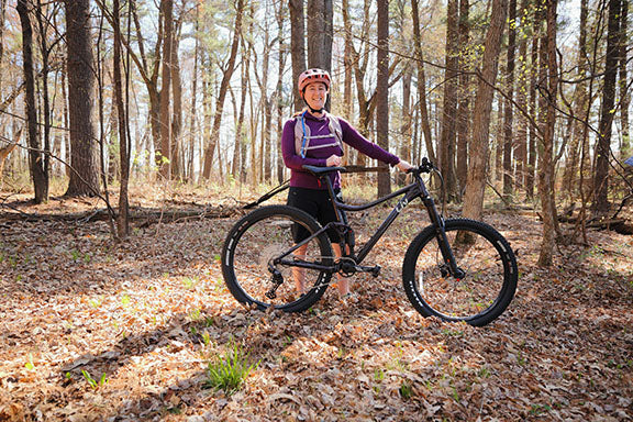A woman stands next to a mountain bike in a wooded setting.