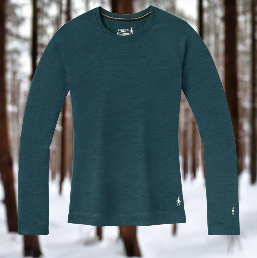 Image of Smartwool women's thermal base layer in heather green with white Smartwool logo on lower left hip. The background is a snow covered winter forest.