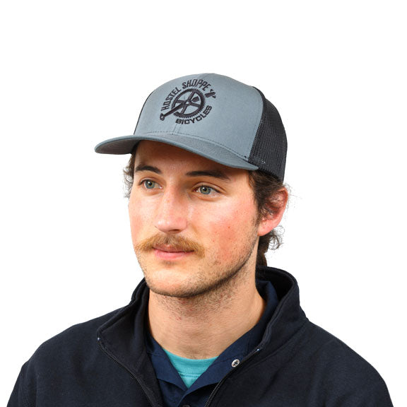 Male model wearing a trucker cap with graphite front panel with black embroidery of Hostel Shoppe logo, and black back half of hat.