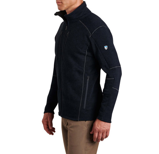 Left profile view of male model wearing a black fleece sweater with a small white and light blue Kuhl logo patch on the left forearm.  