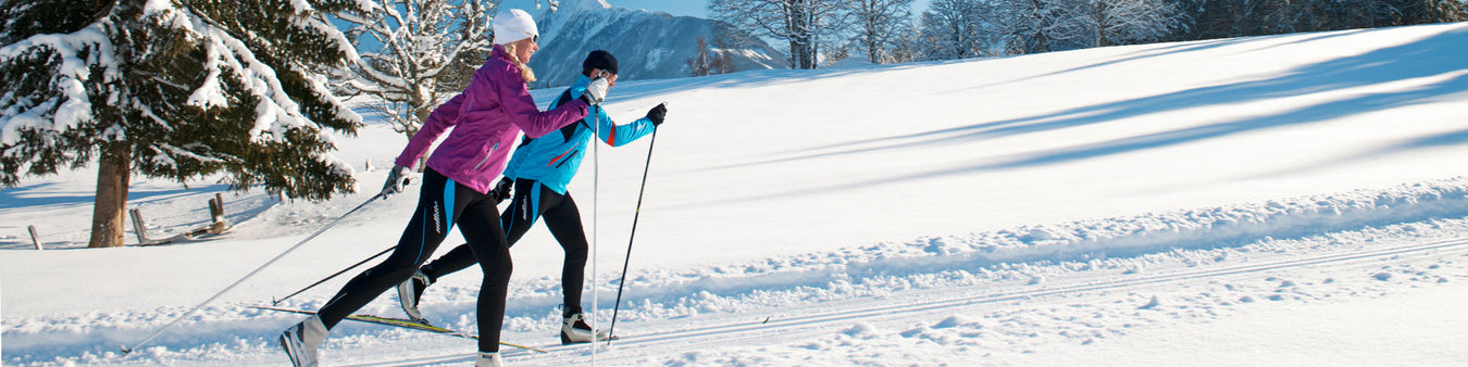 a man and a woman cross country ski thoruh a snowy wooded landscape on a sunny day with mountains in the background.