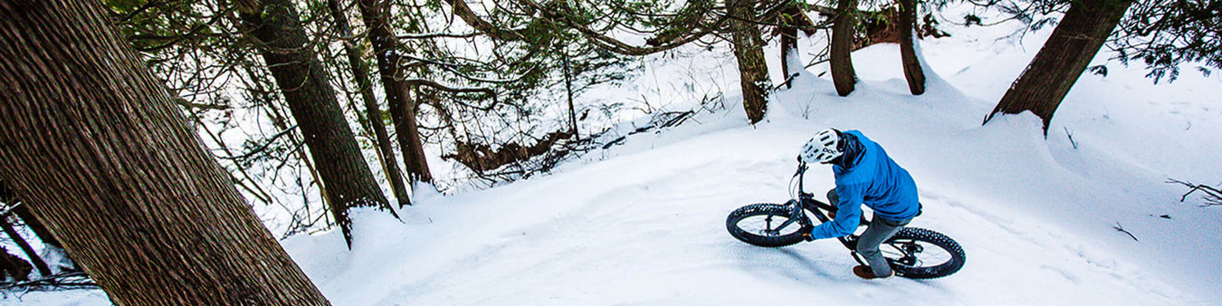 The scene looks down on a rider in a blue jacket riding a black fat bike through the snowy woods.