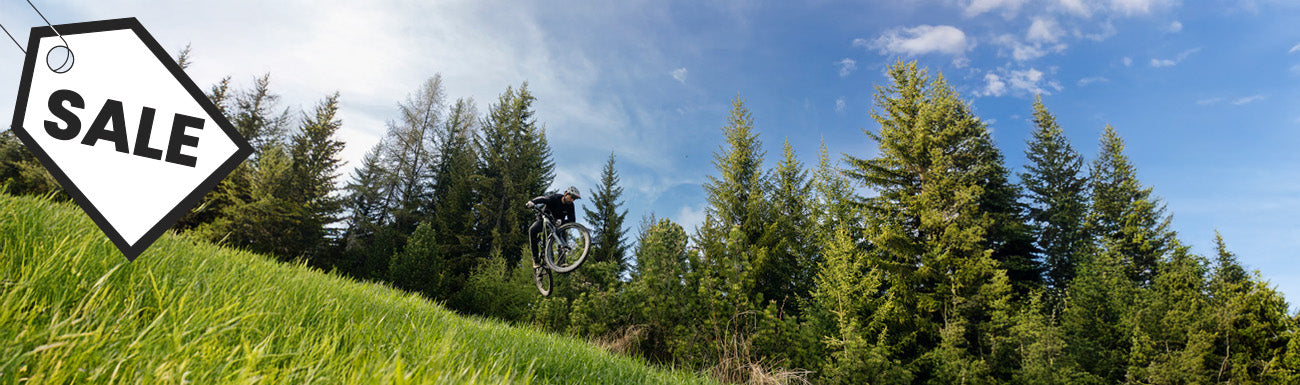 A mountain biker does a jump against a background of blue sky, pine trees and green grass. A black and white "sale" graphic overlays the image.