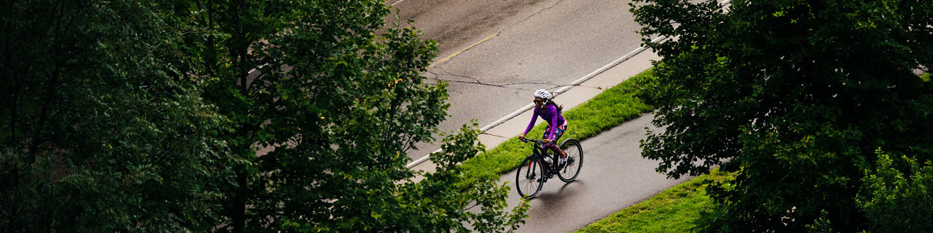 Looking down through the trees, a woman in a purple jersey and flowered tights rides her bike down a bike path that runs alongside the street.