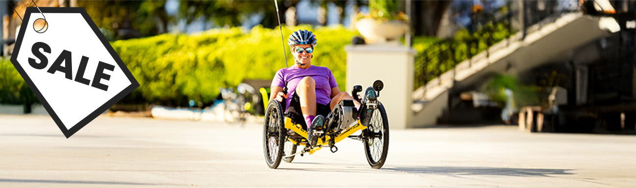 A woman in a purple shirt rides a yellow recumbent trike against a cityscape. A black and white "sale" graphic overlays the image.