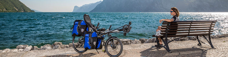 A woman takes a break from riding her recumbent bike, which is parked nearby, on a park bench by a lake surrounded by mountains.