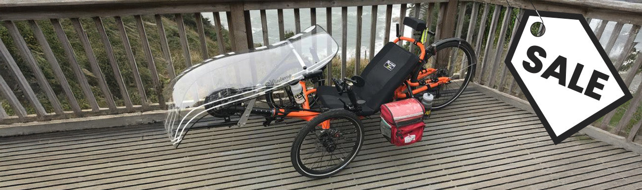 A recumbent trike sits on a deck on a hillside above the ocean and it has all kinds of accessories installed, including a fairing, bag mount and bag, a neckrest and more. A black and white "sale" graphic overlays the image.