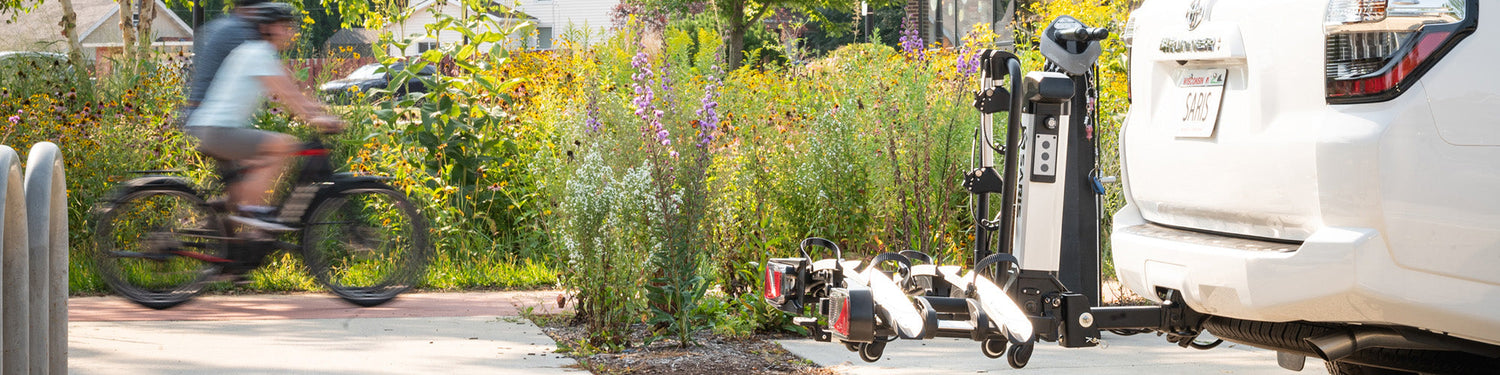 Two bikers ride past a floral background, while a Saris bike hitch rack is shown on a vehicle in the foreground.