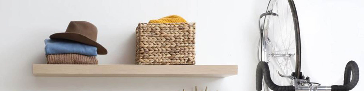 A bicycle hangs from a wall mount hook next  to a shelf with some clothing, a hat and a basket on it.