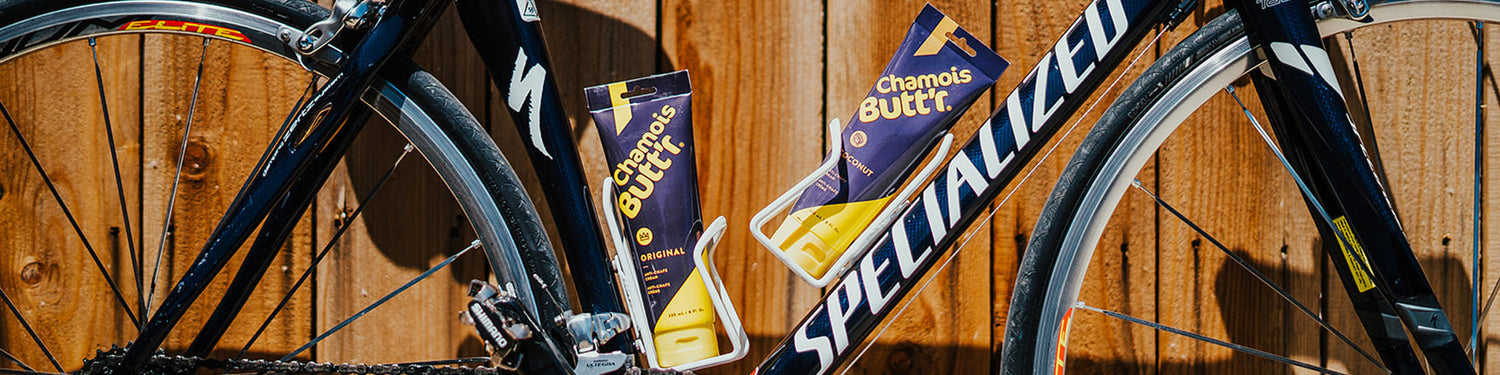 A navy blue Specialized bike with two water bottle cages holding Chamois Butt'r Anti-Chafing Cream against a wooden fence.