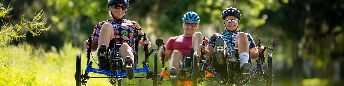 Three recumbent trike riders ride towards the camera  on Catrikes in a wooded setting.