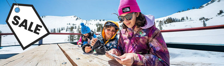 A women and two kids sit at a picnic table in winter clothing with a snowscape in the background. A black and white "sale" graphic overlays the image.