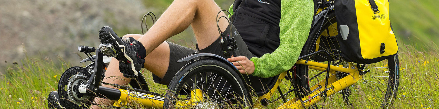 A man in a black and green top with grey shorts rides a yellow Azub trike with fenders on it through a grassy meadow.