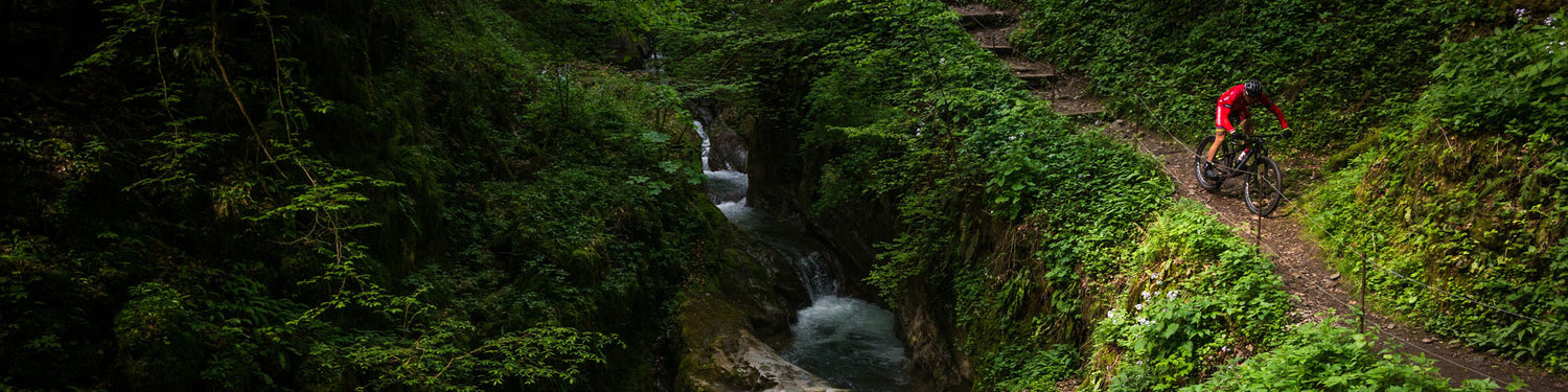 A rider in a bright red outfit rides his mountain bike down a steep trail in the woods alongside a waterfall.