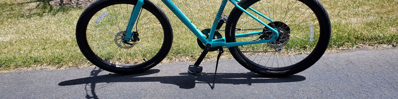 A teal colored bike is propped up with a kickstand on a bike path next to a lawn that is starting to green up in spring.