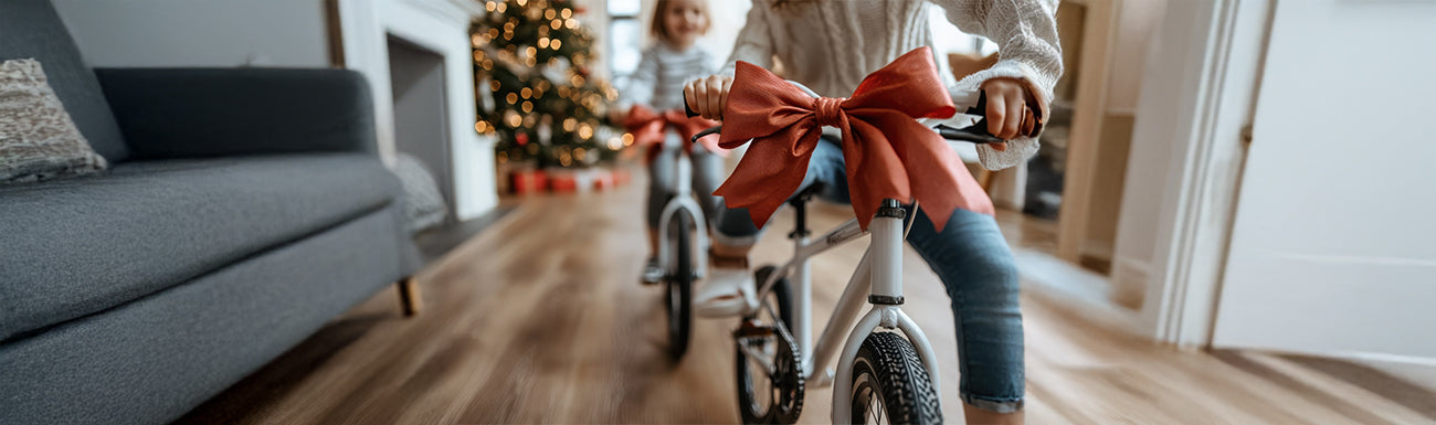 Two girls ride bikes with bows in a living room with a Christmas tree in the background.
