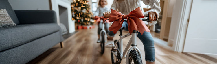 Two girls ride bikes with bows in a living room with a Christmas tree in the background.