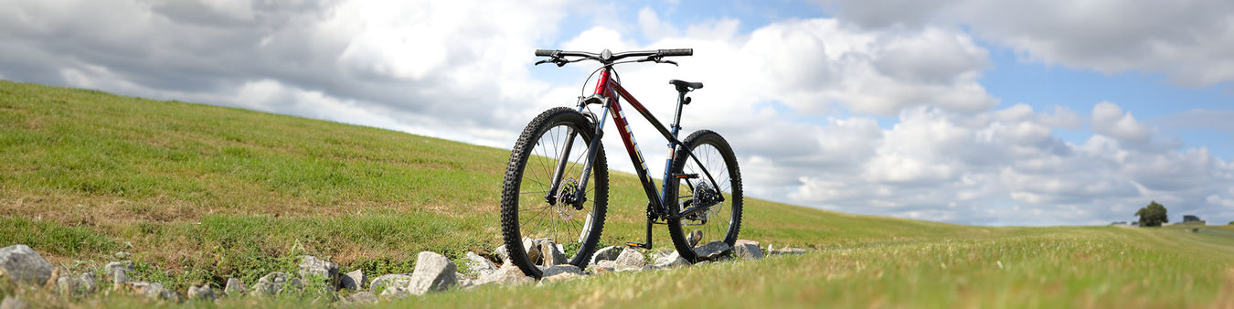 A Trek Marling 6  in red/blue sits amongst some rocks in a grassy landscape under a partly cloudy sky.