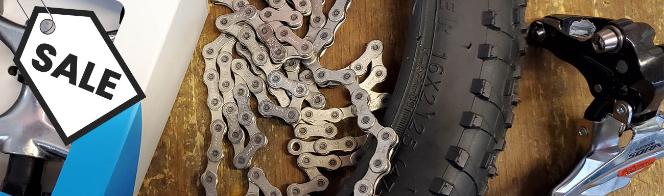 An image of a pedal, chain, tire and front derailleur against a wood backdrop. A black and white "sale" graphic overlays the image.