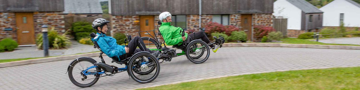 A man and a woman ride recumbent trikes down a brick path with quaint stone and wood buildings in the background.