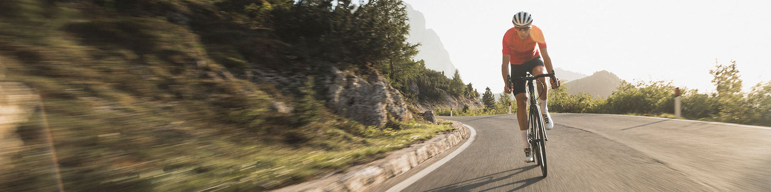 A road cyclist in an orange jersey and black shorts rides his road bike at speed around a corner through mountainous terrain during the day.