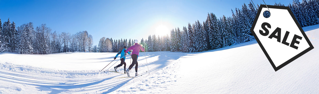 A man and a woman cross country ski across a snowy landscape with trees int he background. A black and white "sale" graphic overlays the image.