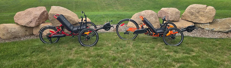 The Catrike Max in red and the Catrike Dumont in orange with e-assist kits installed against a grassy background with landscaping rocks.