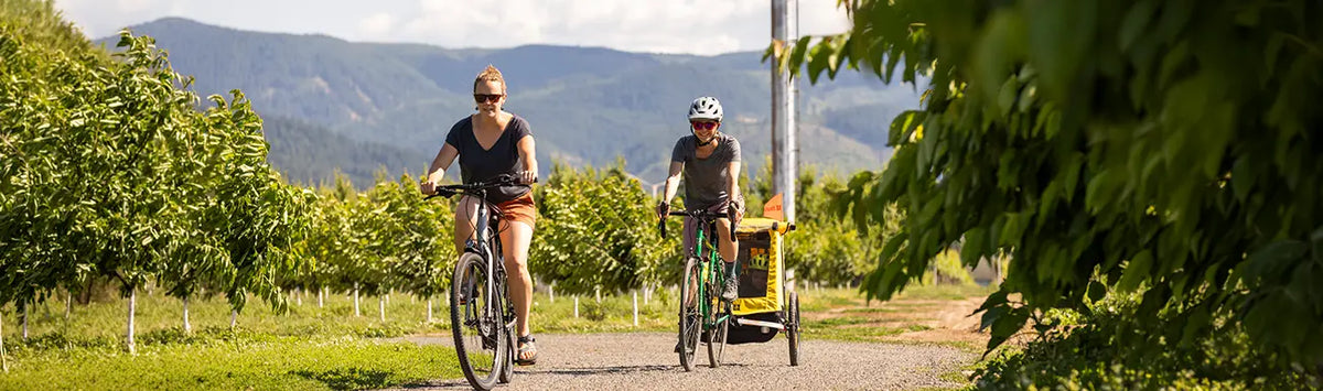 Two women cycle down a country road while one pulls a Burley child trailer on a sunny summer day.