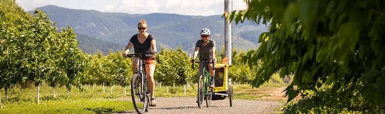 Two women cycle down a country road while one pulls a Burley child trailer on a sunny summer day.