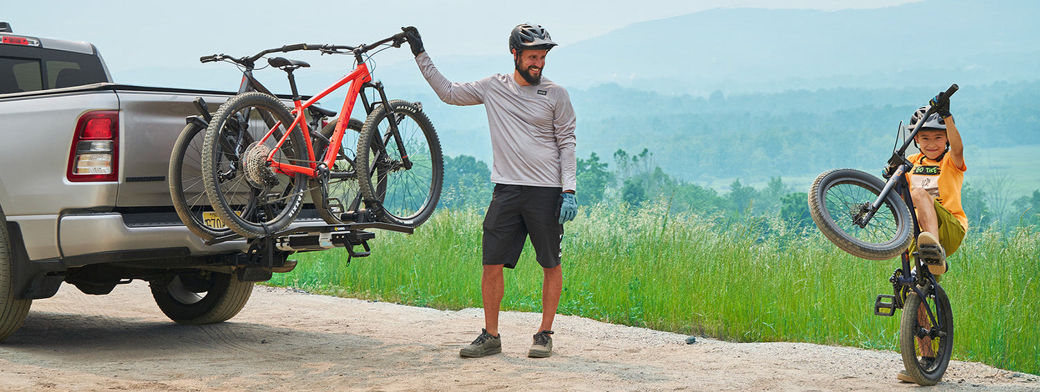 A man stands by a bike rack on his pickup truck, while his son does a wheelie.