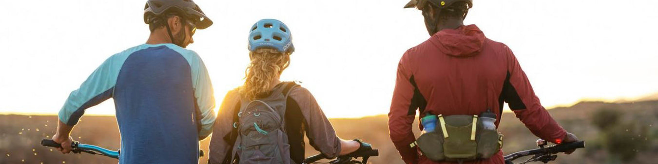 Three cyclists wearing a variety of bike clothing  and helmets walk their bikes towards the sunset.