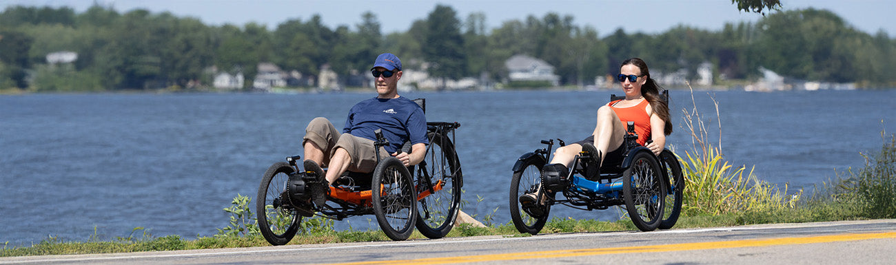 A man and a woman ride Greenspeed Magnum recumbent trikes along a road side near a lake.
