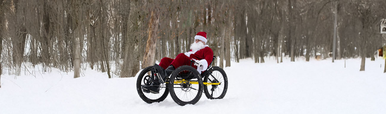 Person dressed as Santa Claus riding a recumbent trike in a snowy forest.