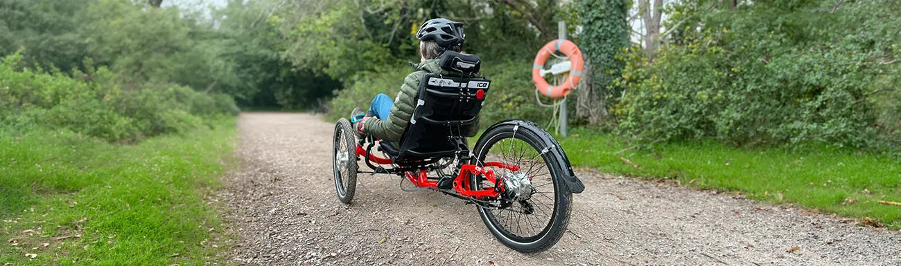 A woman rides an ICE Sprint Pixel recumbent trike down a gravel bike path through the woods.