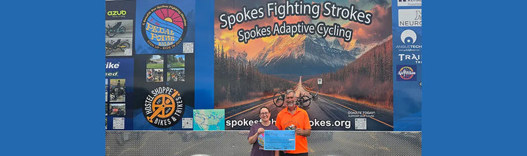 Hostel Shoppe owner, Breanna, and Dan Zimmerman from Spokes Fighting Strokes stand in front of the Spokes Fighting Strokes Trailer, with a fundraising check.