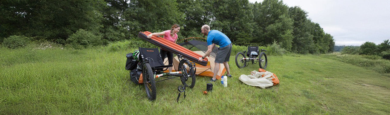 A man and a woman set up camping equipment near two recumbent trikes in a grassy, wooded area.
