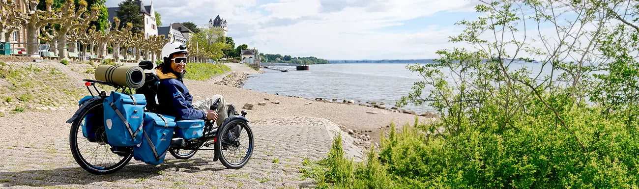 A man on an HP Velotechnik Scorpion trike has racks and panniers for touring, while he takes a break in a town alongside a lake.