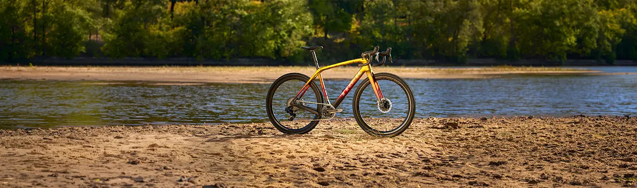 A Trek road bike on a sandy beach along a river with trees in the background.