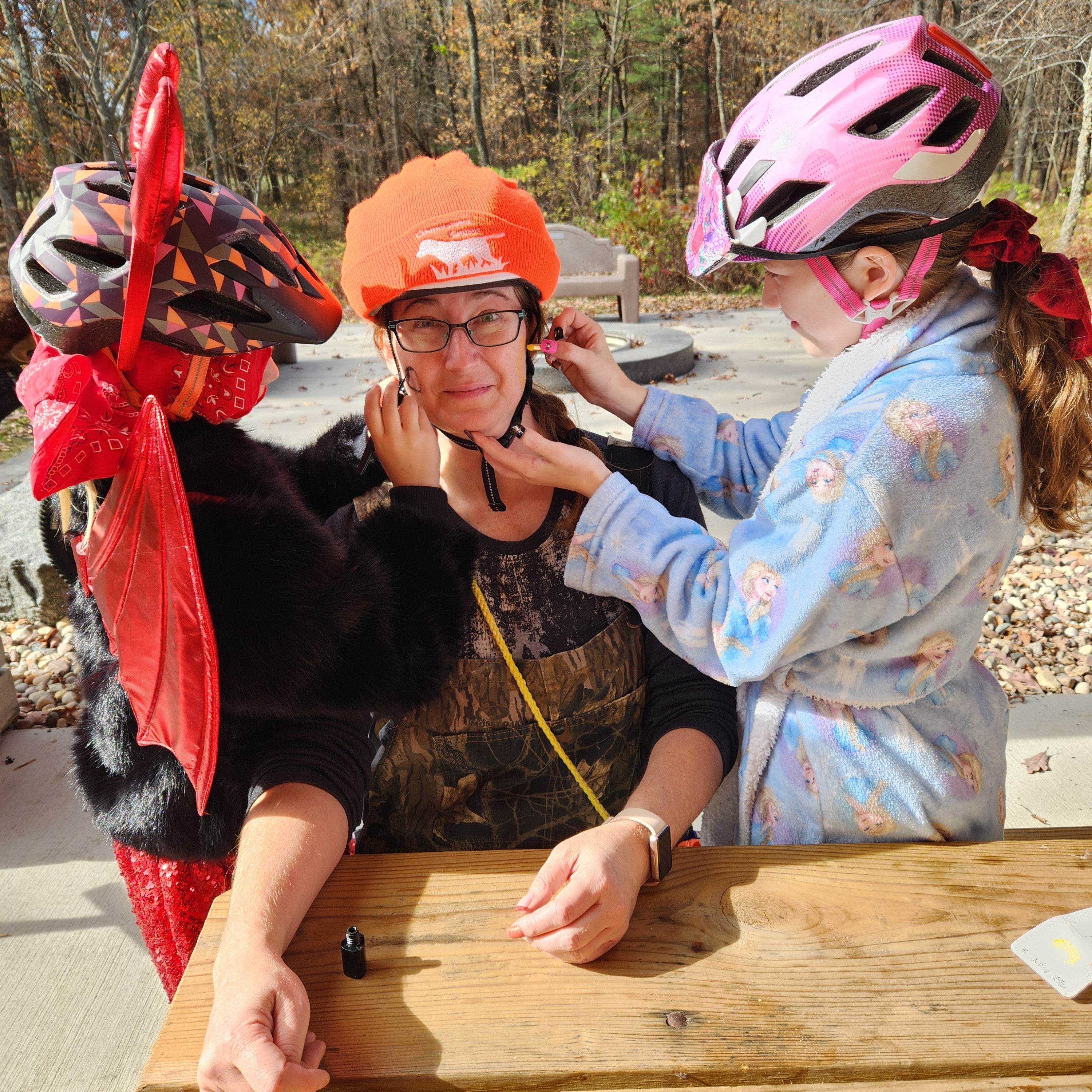 A woman in an orange hat is having her face painted by two girls who are dressed in Halloween costumes with bike helmets on their heads.