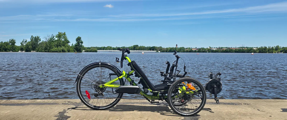 A recumbent electric Catrike with a bright lime green frame and black components is parked on a concrete riverside path with a large body of water and trees in the background under a blue sky.