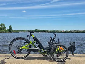 A recumbent electric Catrike with a bright lime green frame and black components is parked on a concrete riverside path with a large body of water and trees in the background under a blue sky.
