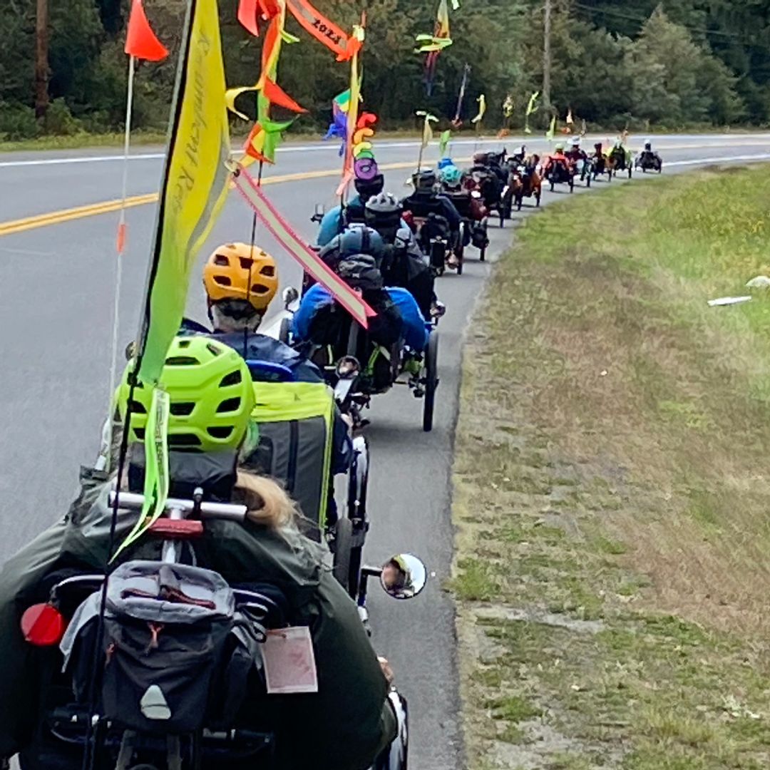 Group of cyclists on trikes with colorful flags on a road surrounded by trees.