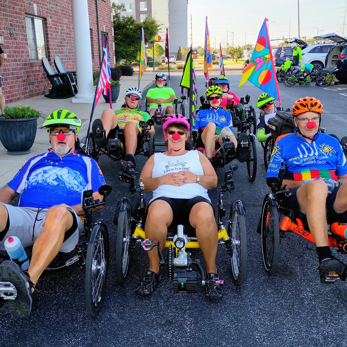 A group of people are sitting on recumbent trikes in a parking lot, wearing red clown noses, bright colorful cycling jerseys, helmets, and sunglasses. Several participants are smiling, and a brick building with white columns is visible on the left.