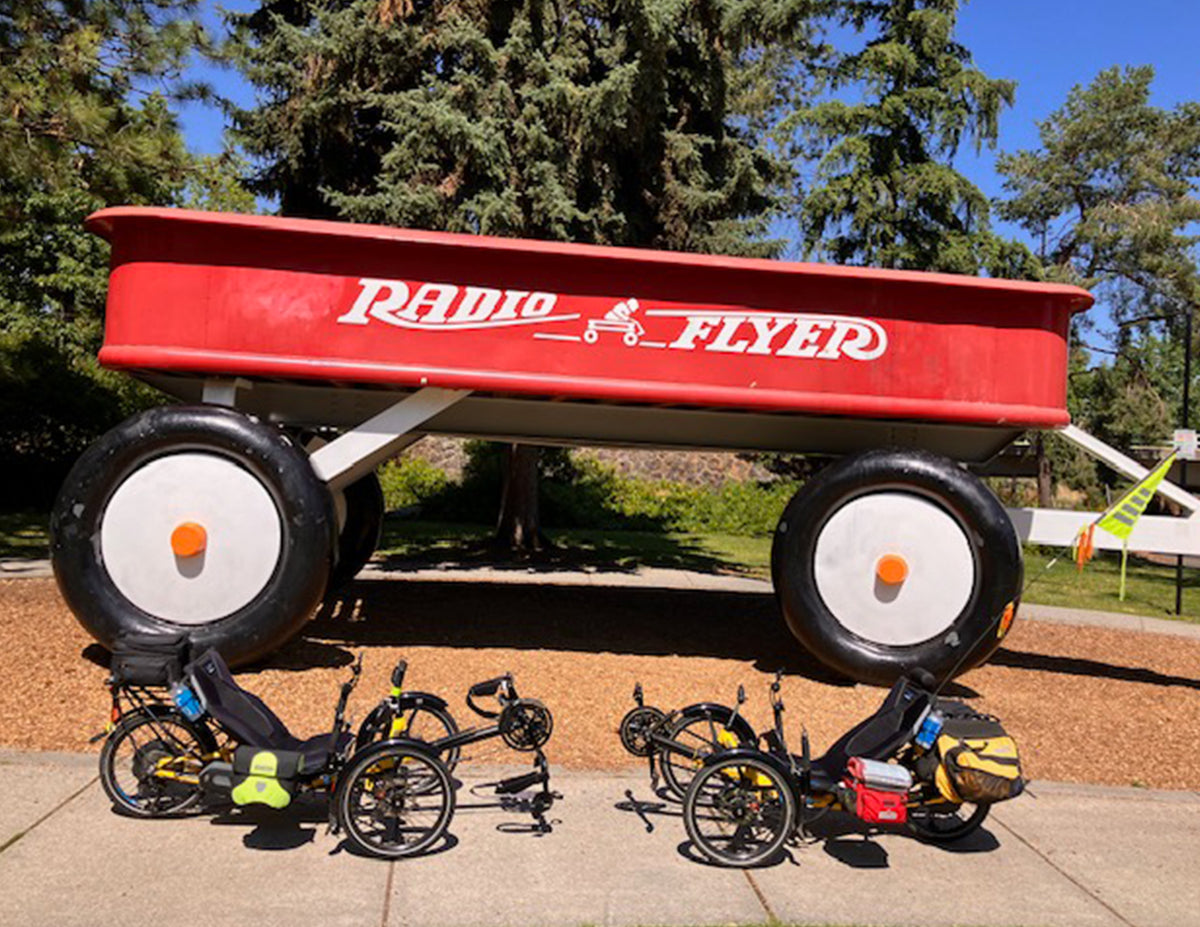 A giant red Radio Flyer wagon displayed outdoors on a paved area, with several black recumbent tricycles parked in front. Trees and blue sky are visible in the background.