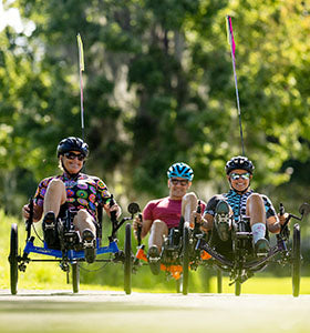 Three people ride Catrike recumbent trikes down a bike path in a wooded area on a sunny day.