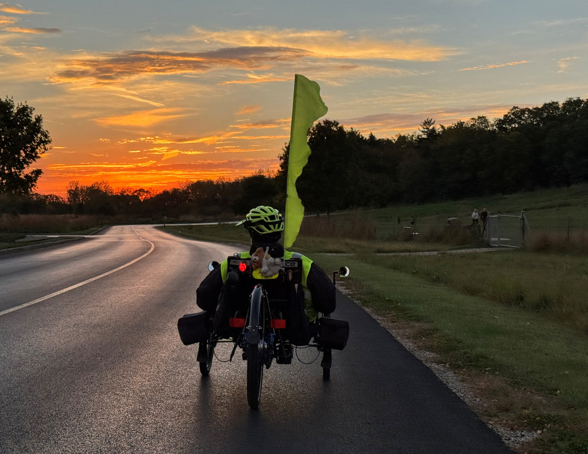 A rear view of a recumbent tricycle with a bright yellow flag and a helmeted rider, riding on a paved road during a vibrant orange and yellow sunset.