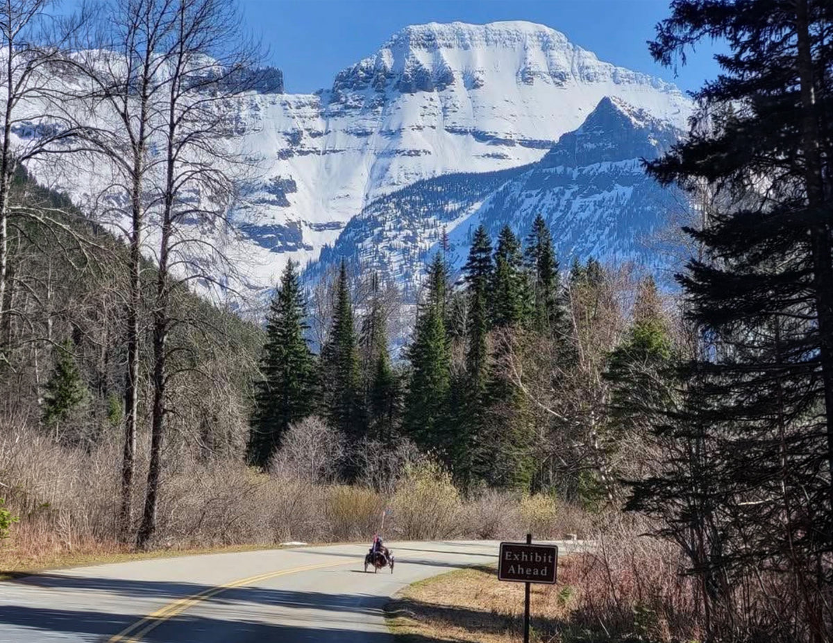 A scenic mountain landscape with a snow-covered peak, evergreen forest, a paved road, and a recumbent triker traveling on the road. A sign on the right reads "Exhibit Ahead".