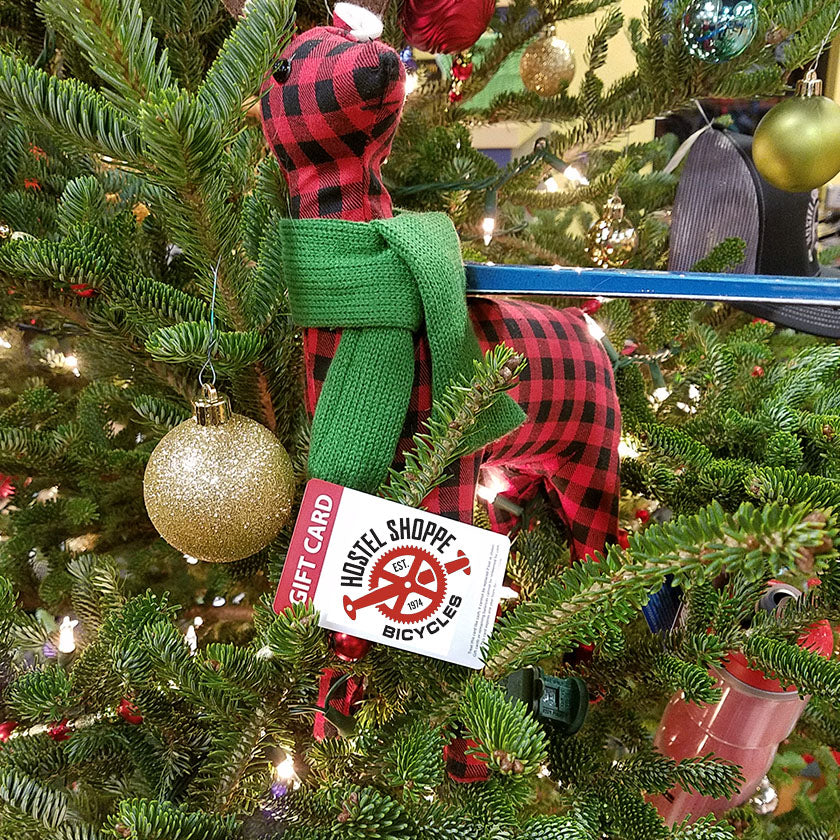 A Hostel Shoppe Gift Card sits in a decorated Christmas tree below a black and red plaid reindeer.